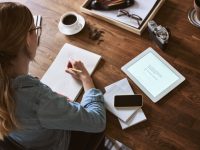 Young woman sitting at a table at home working with a digital tablet and sketching out designs with a pencil on a notepad